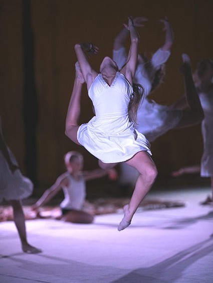 Nora leaping high on stage in a white dress, back arched and arms overhead
